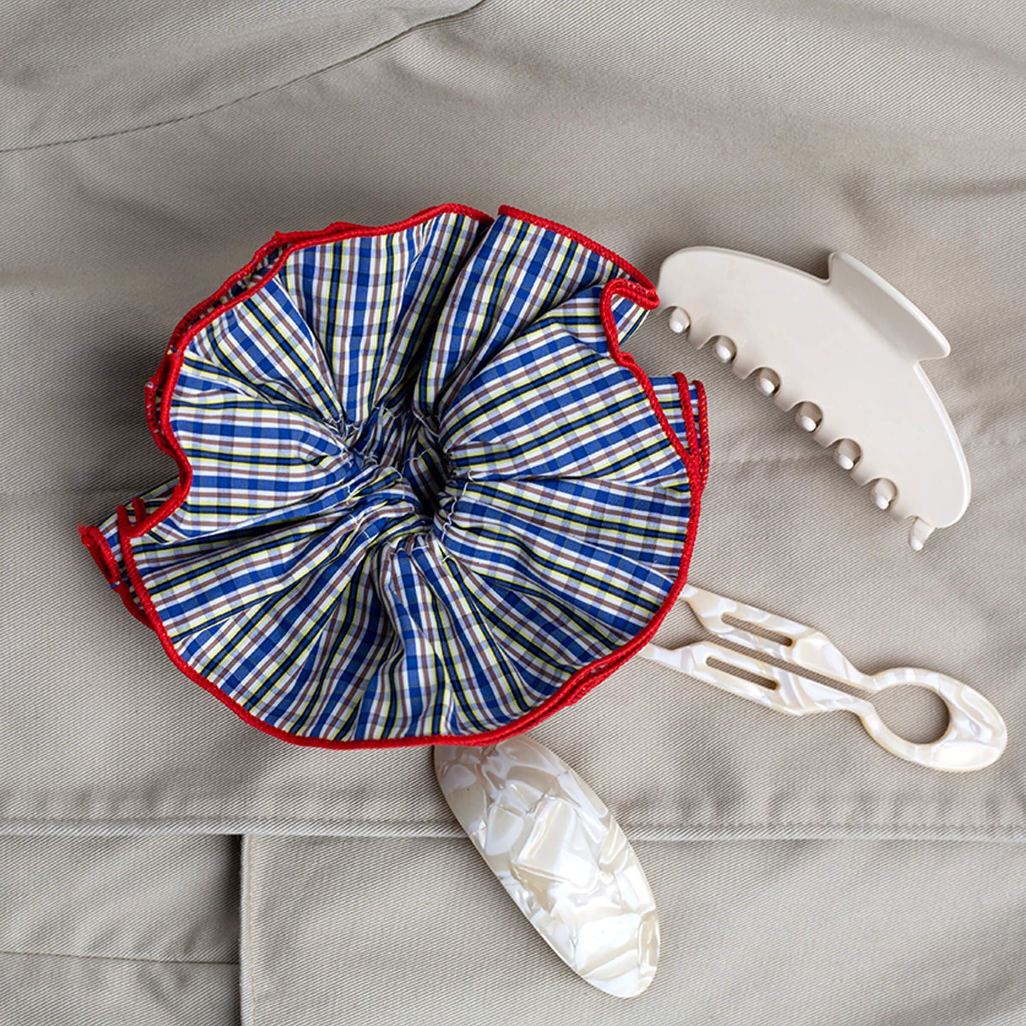 Plaid scrunchie with red trim, next to a cream claw clip, cream hair pin and cream barrette clip on a beige rain coat